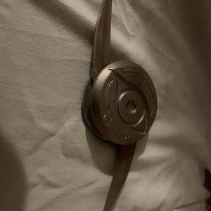 Könnte beinhalten: Ein bronzefarbenes, stilisiertes Objekt mit einem zentralen kreisförmigen Element, das ein Augendesign aufweist. Zwei scharfe, klingenartige Verlängerungen ragen aus dem Mittelteil heraus und erzeugen Symmetrie. Das Objekt scheint aus Metall zu sein.