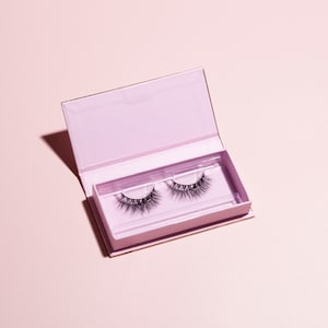 Könnte beinhalten: Ein Paar falsche Wimpern in einer rosa und goldenen Box. Die Wimpern befinden sich in einem durchsichtigen Plastikbehälter und sind bereit zum Auftragen.