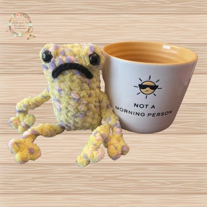 May include: A handmade, yellow and purple crochet frog with a black frown and large black eyes sits next to a white mug. The mug has a yellow interior and reads "NOT A MORNING PERSON" with a sunglasses-wearing sun graphic.