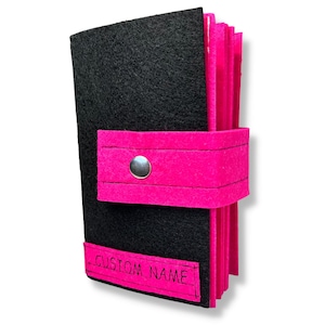 May include: A black felt wallet with a pink felt flap and a silver button. The flap has a pink felt strap with stitching. The wallet has a pink felt bottom and a black felt top. The wallet has a pink felt label with the text "CUSTOM NAME" in black.