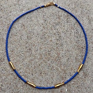 Könnte beinhalten: Eine blaue Perlenkette mit goldfarbenen fischförmigen Anhängern. Die Kette liegt auf einer sandigen Oberfläche, und der Verschluss ist goldfarben. Die Perlen sind klein und gleichmäßig verteilt.