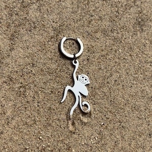 Könnte beinhalten: Ein silberner Ohrring mit einem Affenanhänger. Der Anhänger ist ein stilisierter Affe mit einem lächelnden Gesicht und gebogenen Gliedmaßen. Der Ohrring ist im Creolen-Stil gehalten, und der Affenanhänger hängt an der Creole. Auf sandigem Hintergrund.