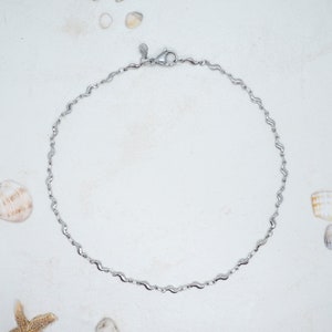 Könnte beinhalten: Ein silbernes Fußkettchen mit wellenförmigem Design. Das Fußkettchen ist aus Metall und hat einen Karabinerverschluss.