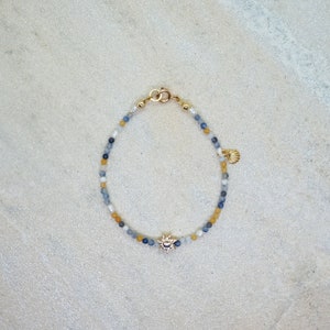 Könnte beinhalten: Ein zartes Armband mit Perlen, einem goldenen Sonnenanhänger und einem Muschel-Anhänger. Das Armband besteht aus blauen, weißen und gelben Perlen.