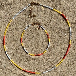 Könnte beinhalten: Ein Perlenkette und Armband-Set. Die Halskette und das Armband bestehen aus kleinen, farbigen Perlen in Gelb, Rot und Grau. Der Schmuck wird auf einer sandigen Oberfläche mit goldfarbenen Verschlüssen präsentiert.