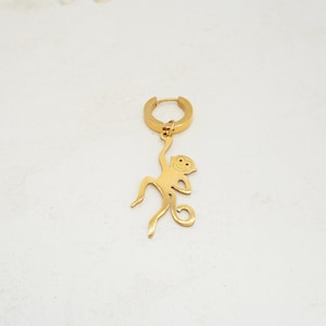 Könnte beinhalten: Goldener Creolenohrring mit einem Affenanhänger. Der Affe hängt mit seinem Schwanz um seinen Arm gewickelt.