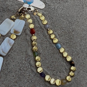 Könnte beinhalten: Eine goldfarbene Halskette mit Muschel- und Edelsteinakzenten. Die Halskette hat abwechselnd muschelförmige Glieder und farbige Edelsteine, darunter rot, grün, blau und lila. Die Halskette wird auf einer sandigen Oberfläche präsentiert.
