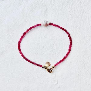 May include: A delicate bracelet featuring small, round, deep red beads. A small, white seashell is centered on the bracelet, with a gold-colored clasp and seashell charm. The bracelet is designed for a delicate, feminine look.