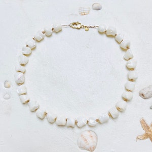 May include: A white shell necklace with irregular, chunky beads. The necklace is strung on a delicate gold chain and features a gold clasp. The background includes seashells and a starfish, suggesting a beach theme.