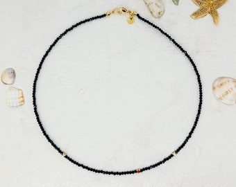 May include: A delicate black beaded necklace with gold accents. The necklace is made of small black beads and features two small gold beads with red stones.
