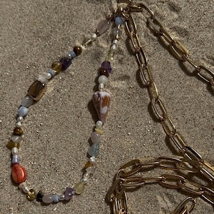 May include: A beaded necklace with a seashell pendant and a gold chain. The necklace features a variety of colorful beads, including brown, purple, and blue, and is set against a sandy background. The gold chain is thick and links together.