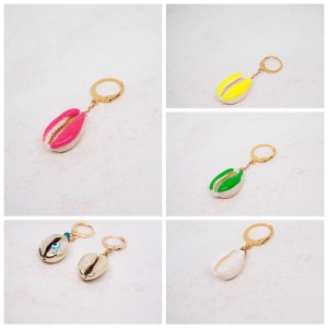 Könnte beinhalten: Fünf verschiedene Schlüsselanhänger mit einem Kaurischnecken-Anhänger. Die Muscheln sind in verschiedenen Farben bemalt, darunter rosa, gelb, grün und weiß. Ein Schlüsselanhänger zeigt eine weiße Kaurischnecke mit einem blauen Glücksbringer-Anhänger.