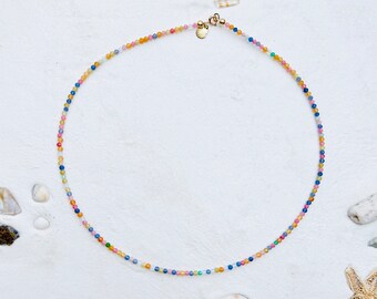 Könnte beinhalten: Eine bunte Perlenkette mit einem Muschel-Anhänger. Die Kette besteht aus kleinen, runden Perlen in Blautönen, Rosa, Gelb und Grün. Die Perlen sind auf einer dünnen goldenen Kette aufgefädelt.