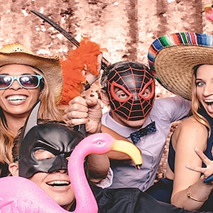 Puede incluir: Un grupo de amigos con sombreros de fiesta y m&aacute;scaras posan para un photocall. Sostienen varios accesorios, incluyendo un flamenco rosa, una maraca y una boa de plumas. El fondo es un tel&oacute;n de fondo dorado brillante.