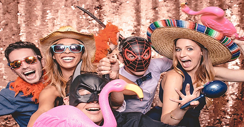 Puede incluir: Un grupo de amigos con sombreros de fiesta y m&aacute;scaras posan para un photocall. Sostienen varios accesorios, incluyendo un flamenco rosa, una maraca y una boa de plumas. El fondo es un tel&oacute;n de fondo dorado brillante.