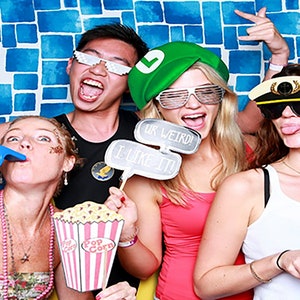 Puede incluir: Cuatro amigos posan para una foto de cabina fotogr&aacute;fica. Llevan sombreros y gafas divertidas. Una persona sostiene un cartel que dice "Ur Weird! I Like It!" Otra persona sostiene un cubo de palomitas de ma&iacute;z. El fondo es una pared de azulejos azules y blancos.