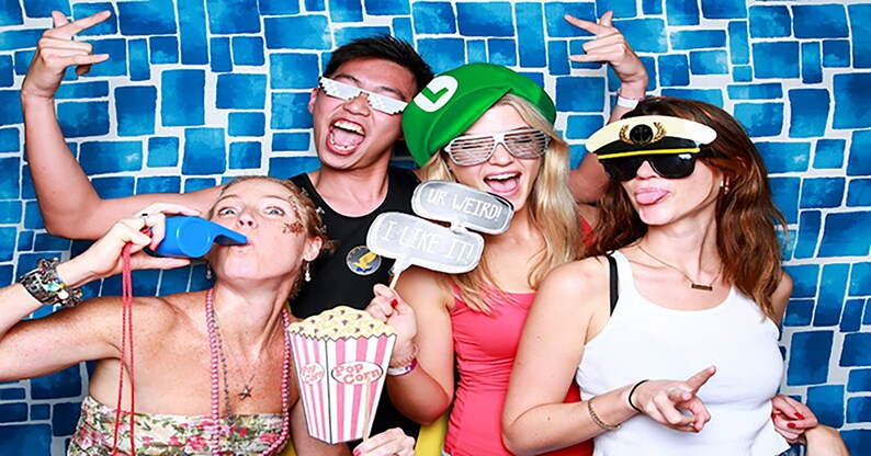 Puede incluir: Cuatro amigos posan para una foto de cabina fotogr&aacute;fica. Llevan sombreros y gafas divertidas. Una persona sostiene un cartel que dice "Ur Weird! I Like It!" Otra persona sostiene un cubo de palomitas de ma&iacute;z. El fondo es una pared de azulejos azules y blancos.