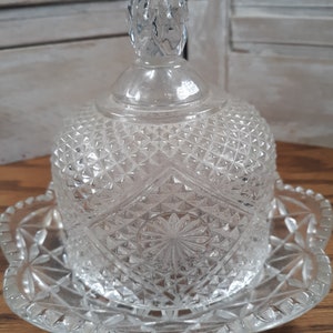 May include: A clear glass butter dish with a domed lid and a scalloped base. The lid has a knob on top and the dish has a diamond pattern.