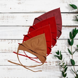 Peut inclure: Quatre bonnets pour bébés de différentes nuances de rouge et de marron sont disposés en diagonale sur une surface en bois blanc. Les bonnets ont une forme triangulaire avec des liens. Des feuilles vertes sont en arrière-plan.