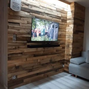 Può includere: Una parete rivestita con assi di legno di recupero orizzontali con un televisore a schermo piatto montato e una soundbar nera. Un orologio da parete decorativo bianco con il testo SPRAW SZANSE STAC SIE ZYCIA è montato in alto a sinistra.