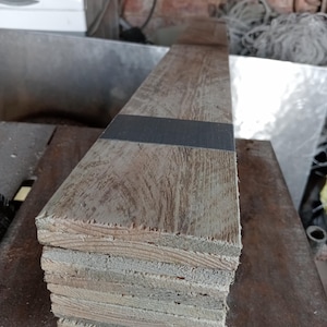 May include: A stack of six rectangular wooden boards with a weathered, light brown finish and visible grain patterns. A single, solid black rectangular strip is positioned horizontally across the top board, creating a contrast against the wood.