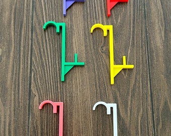 May include: A collection of six colorful plastic curtain rod holders in purple, red, green, yellow, pink, and white. The holders are arranged on a brown wooden surface. Each holder has a unique shape with a curved top and a vertical support.