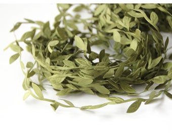 Puede incluir: Un rollo de guirnalda de hojas verdes, ideal para manualidades o decoración. La guirnalda presenta pequeñas hojas espaciadas uniformemente en un tono verde apagado. Las hojas están unidas a un tallo delgado y flexible. Fondo blanco.