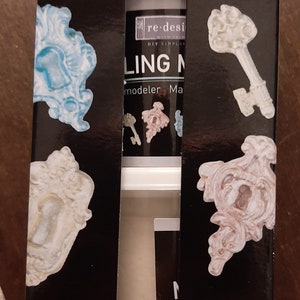 Puede incluir: Una caja negra de material de modelado con el texto "MODELING MATERIAL" en blanco. La caja contiene dos botes de material de modelado no tóxico, cada uno de 100g. Se muestran diseños decorativos de llaves y marcos.