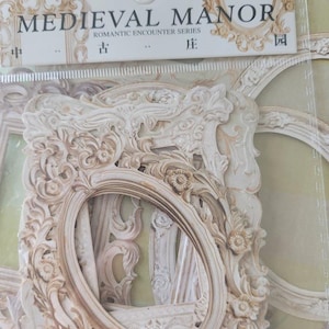 Peut inclure: Un ensemble de 10 cadres en papier décoratifs blancs avec des motifs ornés. Les cadres sont de formes et de tailles variées, et ils sont tous de style vintage. L'emballage indique "MEDIEVAL MANOR ROMANTIC ENCOUNTER SERIES".