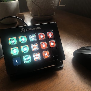 Puede incluir: Un Stream Deck negro con 15 botones, cada uno con una etiqueta naranja y verde diferente. Las etiquetas incluyen "Buy LMT", "Buy 5 LMT", "Target", "Sell LMT", "Sell 5 LMT", "Buy 1/2", "Buy 50x", "Stop Loss", "Sell 1/2", "Sell 10x", "Buy Order", "Cancel Orders", "Sell Orders", y "<" . El Stream Deck está sobre una superficie de madera.