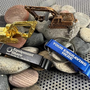 Könnte beinhalten: Drei Schlüsselanhänger mit einem goldenen, kupfernen und blauen Flaschenöffner. Der Flaschenöffner trägt den Text "Your Engraving Here". Die goldenen und kupfernen Schlüsselanhänger zeigen Miniaturmodelle von Baggern. Der goldene Schlüsselanhänger trägt den Text "Equipment Share Grand Junction, CO 970.665.2083".