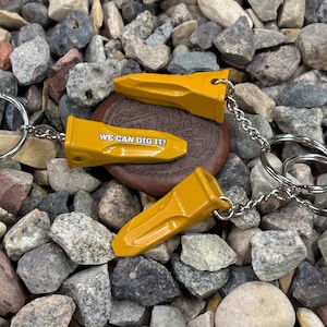 Puede incluir: Llaveros metálicos amarillos con forma de dientes de excavadora, con el texto "WE CAN DIG IT!" en uno. Los dijes están unidos a llaveros y cadenas plateadas, descansando sobre un lecho de rocas grises y marrones.