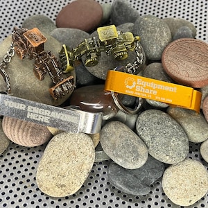 Könnte beinhalten: Zwei Flaschenöffner aus Metall mit Schlüsselanhängern. Ein Öffner ist silberfarben mit dem Text "YOUR ENGRAVING HERE". Der andere Öffner ist golden mit dem Text "Equipment Share" und einem Logo. Beide Öffner sind von kleinen grauen Steinen umgeben.