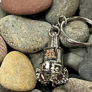 May include: Silver metal keychain shaped like a drill bit with the text "RIG 235" engraved on the side. The keychain is on a bed of gray and brown rocks.