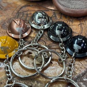 Puede incluir: Llaveros en miniatura con forma de casco de obra en cobre, plata, negro y amarillo. Cada casco presenta una cadena y un anillo plateados, con una pequeña bandera americana y el texto "RIC 521".