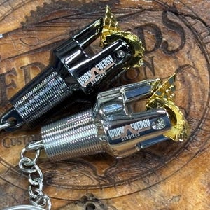 May include: Two metal keychains, one black and one silver, each featuring a detailed drill bit design with gold-coloured accents. The keychains have the text "WORK ENERGY SERVICES" on them and are attached to a silver chain.