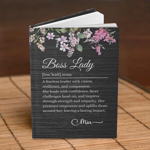 May include: A black journal with a floral design and the text "Boss Lady [bos 'lerdi] noun A fearless leader with vision, resilience, and compassion. She leads with confidence, faces challenges head-on, and inspires through strength and empathy. Her presence empowers and uplifts those around her, leaving a lasting impact. Mia"