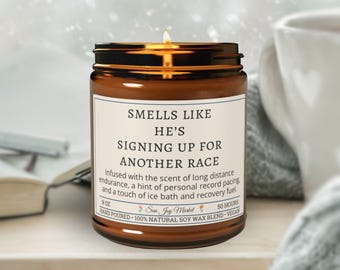 May include: A lit, brown glass jar candle with a gold lid. The label reads "Smells Like He's Signing Up For Another Race." The candle is 9 oz and made with natural soy wax. The candle is next to a white mug.