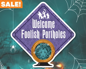 May include: A purple and white diamond-shaped sign with the text "Welcome Foolish Mortals" and a round, gold-rimmed porthole with a green-skinned woman with long, dark hair looking out. The sign is on a dark green background with white spiderwebs.