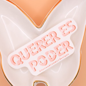 Op de afbeelding: Roze en witte sticker met de tekst "QUERER ES PODER" in een gestileerd lettertype. De sticker zit op een wit keramiek vlinder-schoteltje met een gouden rand. De tekst "@latindreamerdesigns" staat in de rechterbenedenhoek van de sticker.