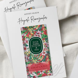 May include: A white card with the text "Afiyet Olsun Hayırlı Ramazanlar" in black calligraphy. The card features a small packet of Japanese Cherry Blossom scented cologne tissues with the brand name "Eyüp Sabrı Tuncer" and the year "1923" printed on it.