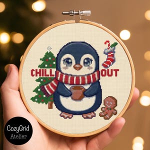 May include: A cross-stitch embroidery in a round wooden hoop featuring a penguin wearing a red and white striped scarf, holding a brown mug. The design includes a green Christmas tree, a red and white stocking, and a gingerbread man with the text CHILL OUT.