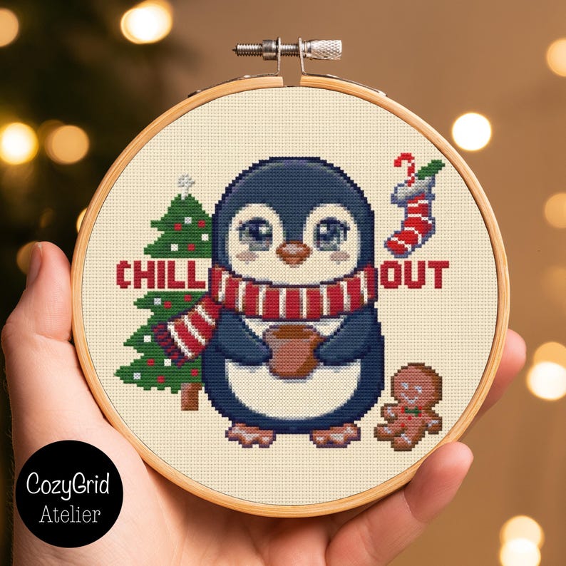 May include: A cross-stitch embroidery in a round wooden hoop featuring a penguin wearing a red and white striped scarf, holding a brown mug. The design includes a green Christmas tree, a red and white stocking, and a gingerbread man with the text CHILL OUT.