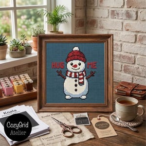 May include: A framed cross-stitch embroidery featuring a white snowman wearing a red knit hat and a red and white striped scarf. The snowman is centered on a blue background with the red text HUG ME. The frame is dark brown wood.
