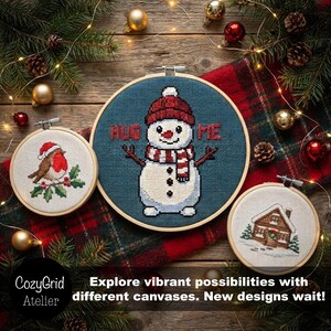 May include: Three circular embroidery hoops featuring cross-stitch designs: a robin on a branch, a snowman wearing a red hat and scarf with the text HUG ME, and a small house in the snow. The hoops are arranged on a red plaid fabric.