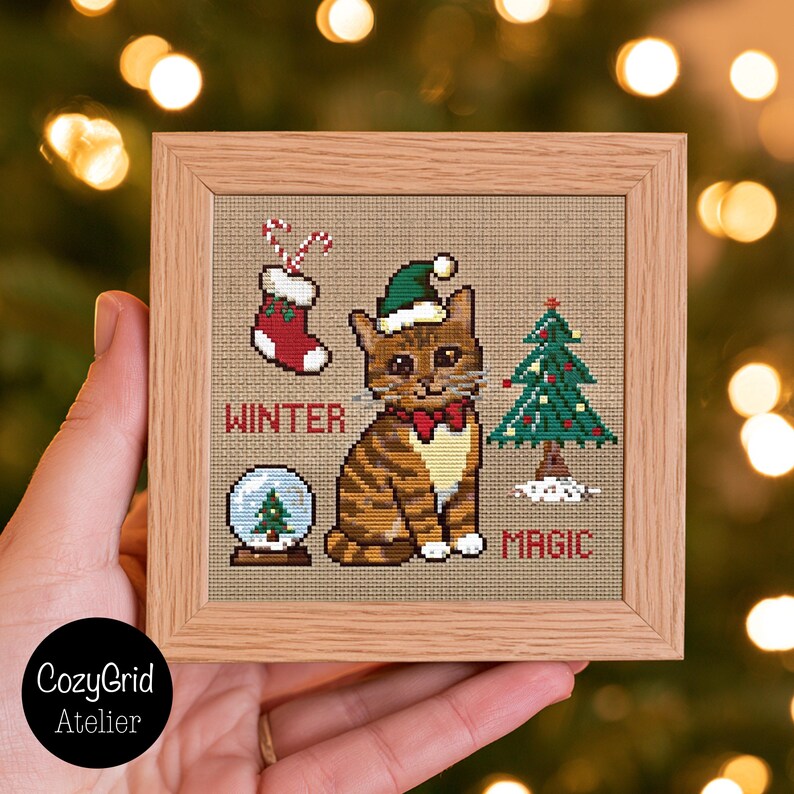 K&ouml;nnte beinhalten: Ein quadratischer Holzrahmen mit einer Kreuzstichstickerei auf beigem Stoff. Das Motiv zeigt eine braune getigerte Katze mit gr&uuml;ner Weihnachtsm&uuml;tze und roter Schleife, einen roten Strumpf, eine Schneekugel und einen Weihnachtsbaum mit dem Text WINTER MAGIC.
