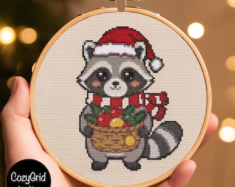 May include: A cross-stitch embroidery in a round wooden hoop featuring a gray raccoon wearing a red and white Santa hat and a red and white striped scarf. The raccoon holds a brown basket filled with red and yellow items. CozyGrid Atelier.