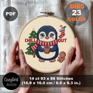 May include: A cross-stitch pattern in a wooden hoop featuring a penguin wearing a red and white striped scarf, holding a brown mug. The design includes a green Christmas tree, a red and white stocking, a gingerbread man, and the text CHILL OUT.