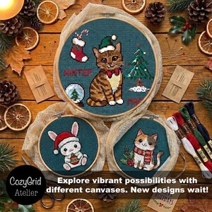 K&ouml;nnte beinhalten: Drei runde Holzstickrahmen mit Kreuzstichmotiven auf dunkelblauem Stoff. Der gr&ouml;&szlig;te Rahmen zeigt eine getigerte Katze mit Weihnachtsm&uuml;tze, einen Weihnachtsbaum, einen Strumpf und eine Schneekugel. Zwei kleinere Rahmen zeigen einen Hasen und eine Katze.