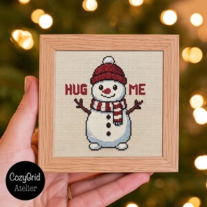 May include: A square cross-stitch embroidery in a light wood frame featuring a white snowman wearing a red knit hat and a red and white striped scarf. The snowman has stick arms, three black buttons, and the text HUG ME in red.